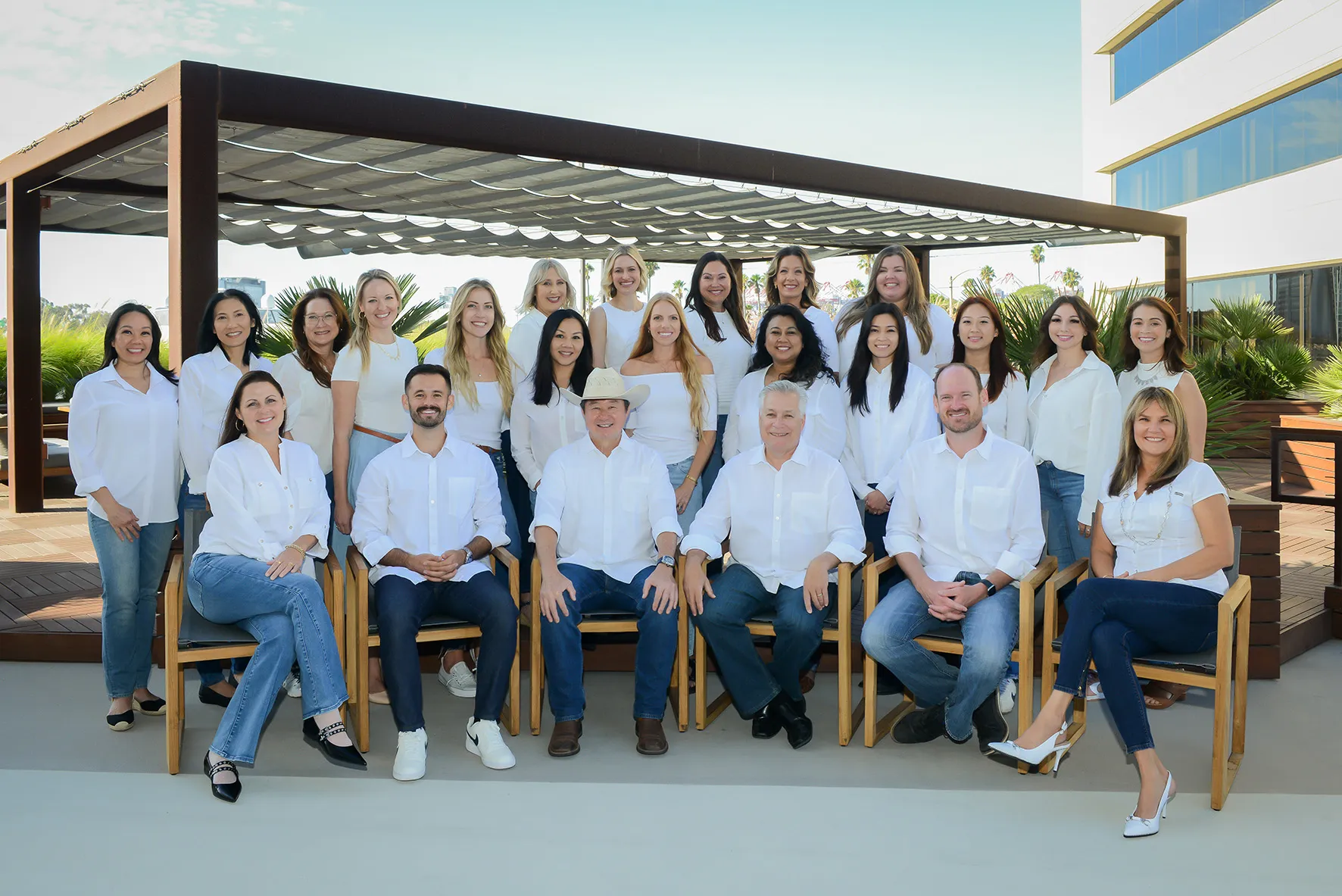 Diverse team of 16 staff members (12 women, 4 men) in coordinated white tops and jeans or dark pants, seated and standing under a modern pergola with greenery — smiling for a group portrait