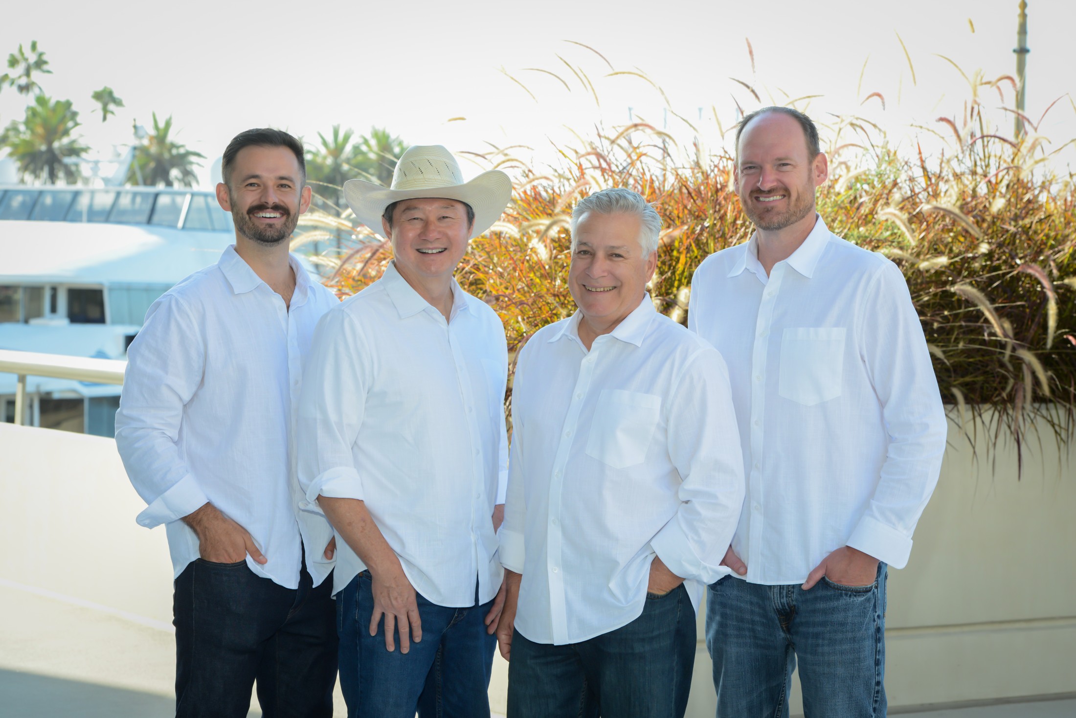 Four male clinicians or leaders in white button-down shirts and dark pants, standing side-by-side on a rooftop terrace with palm trees, blue sky, and a building in the background